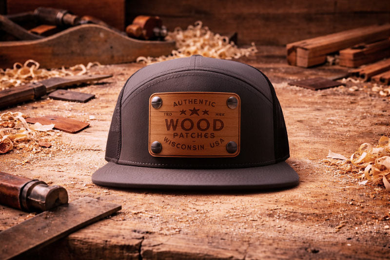 Charcoal 7 panel, flat brim trucker cap with wood patch on a wooden workbench surface with tools and wood shavings